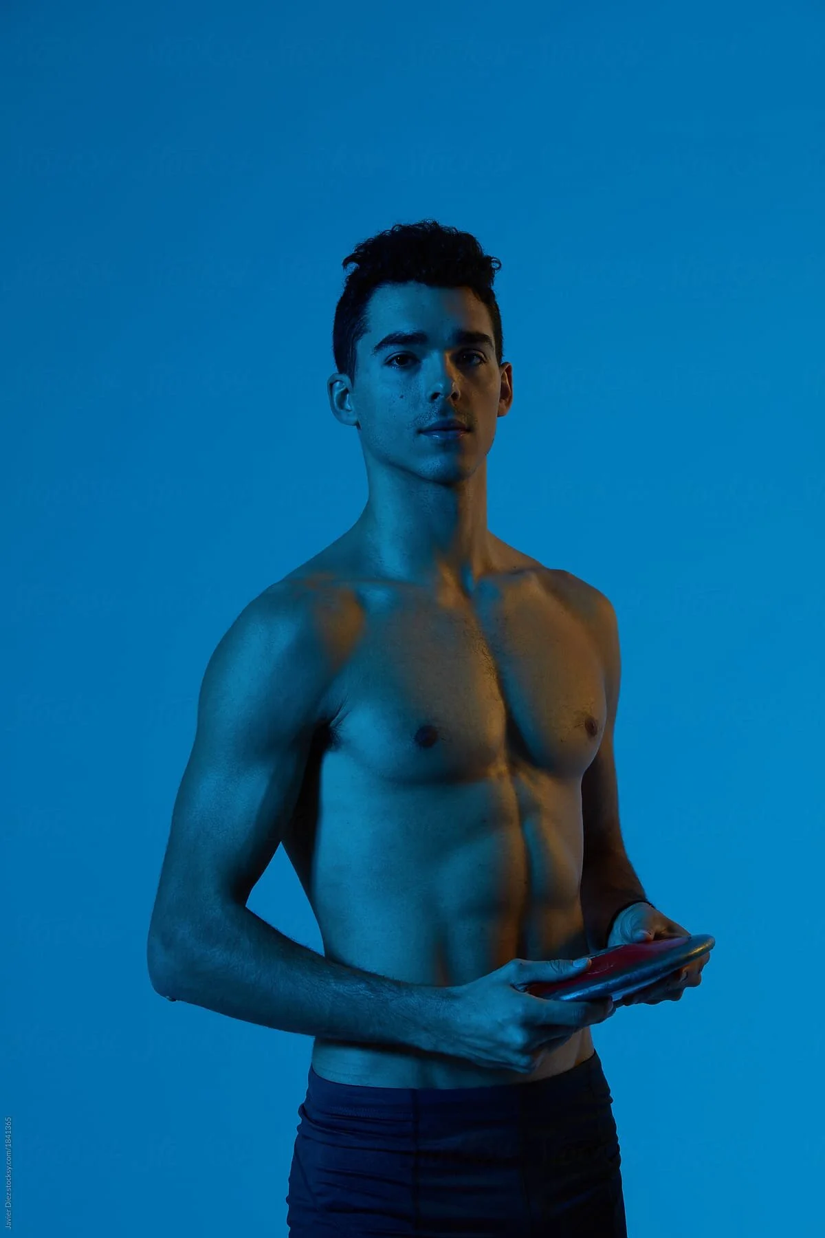 Portrait of young shirtless man with muscular body holding disc and looking at camera in blue light.