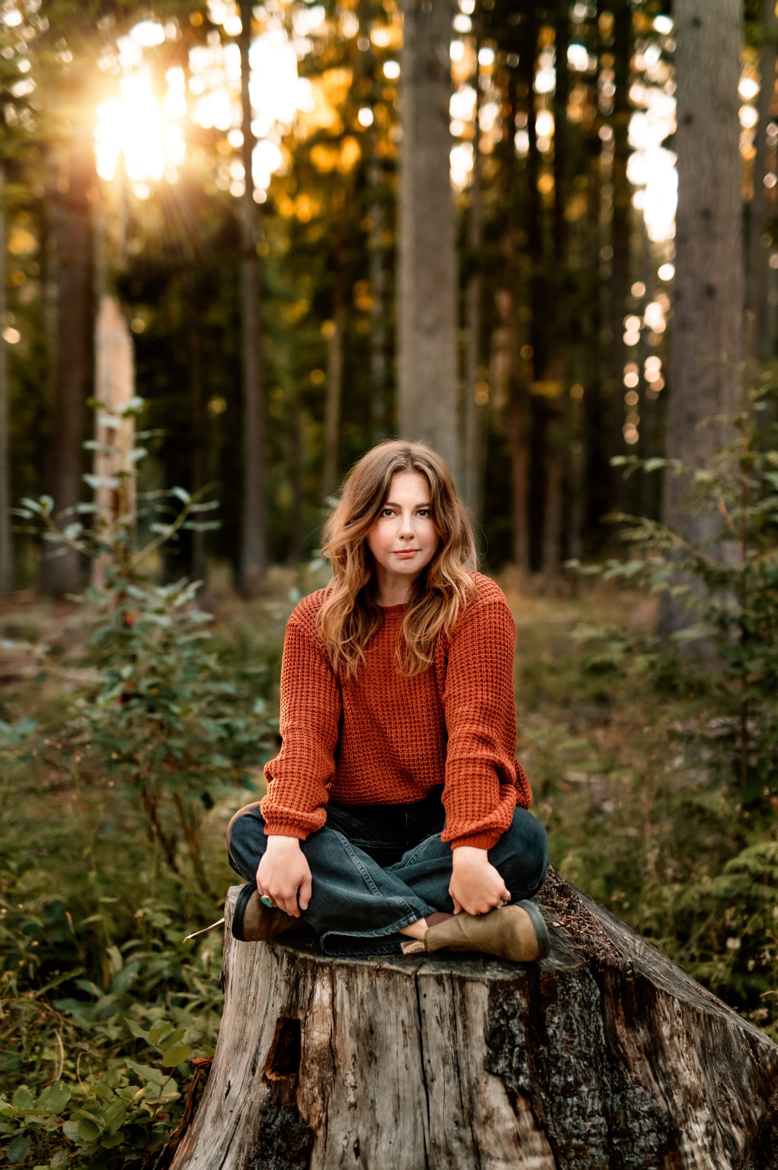 A woman with wavy brown hair sits cross-legged on a tree stump in a forest during sunset, wearing a rust-colored sweater and blue jeans.