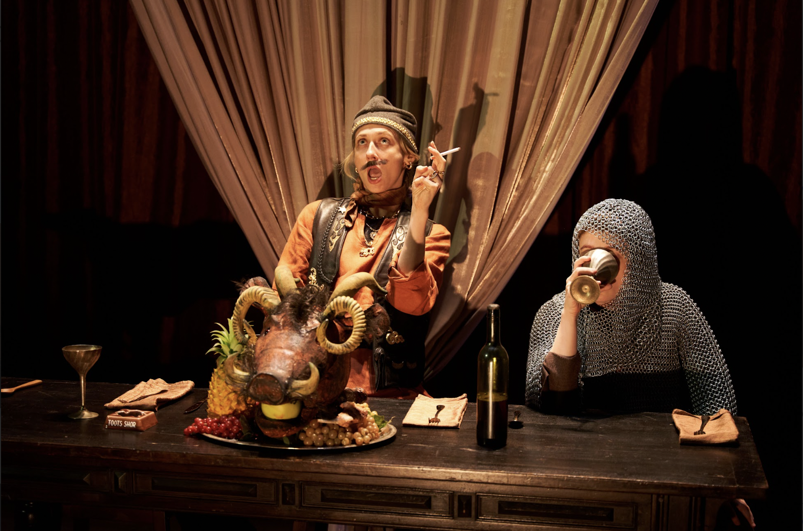 An actor holding a cigarette sits at a table in front of a roasted boar's head next to an actor wearing chainmail armor and drinking from a chalice