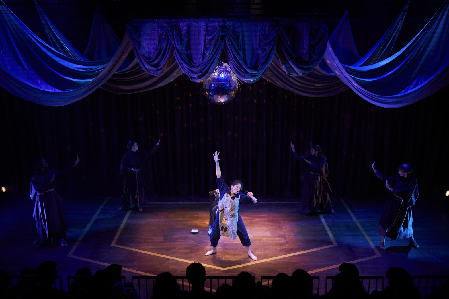 An actor singing into a microphone with one hand in the air under a disco ball surrounded by actors on the edges of the stage holding their arms up