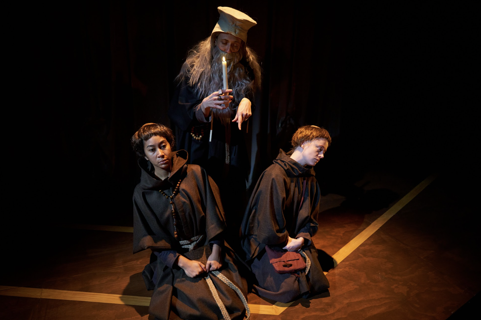 Two actors dressed in hermit's clothing kneel on either side of an actor wearing a long gray beard and holding a candle