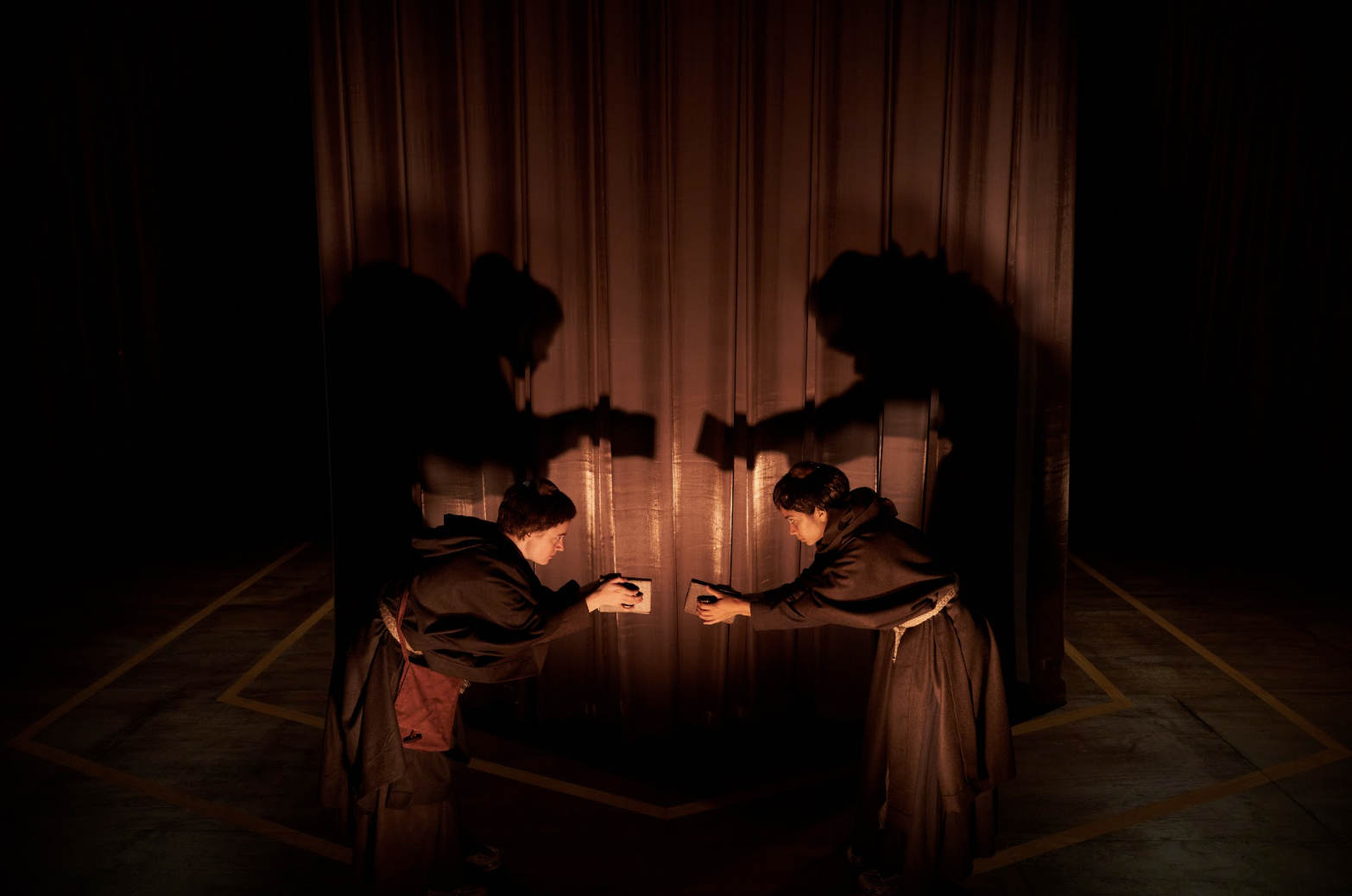 Two actors dressed in hermit's clothing bowing to each other with books in their hands 