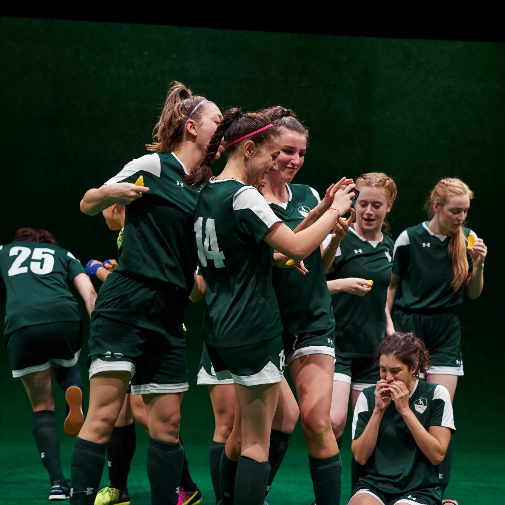 A group of people in green and white soccer uniforms eating orange slices and looking at something together on a cell phone