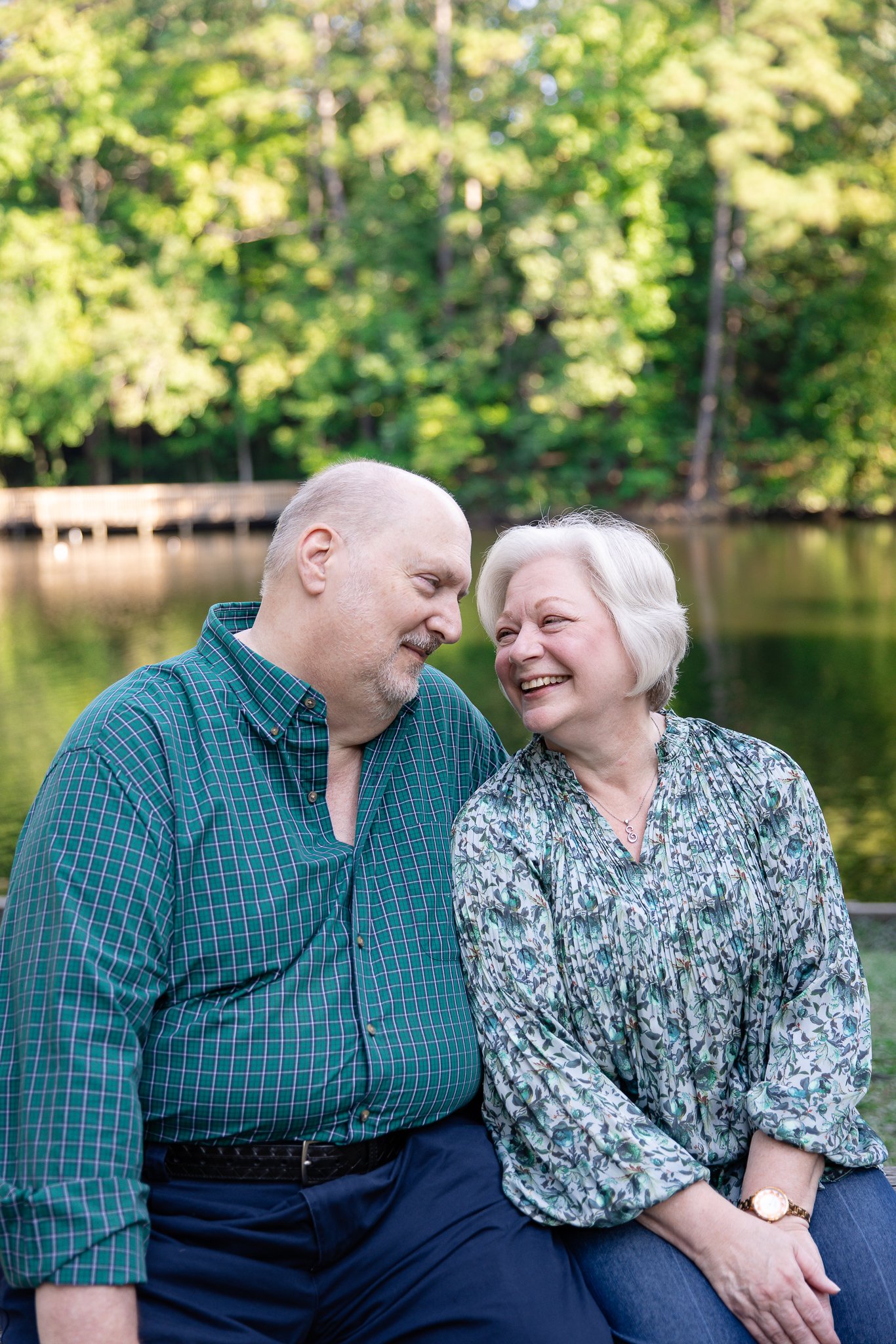 An older couple sitting on a bench outdoors near a body of water, smiling and looking at each other, with trees and a wooden dock in the background.