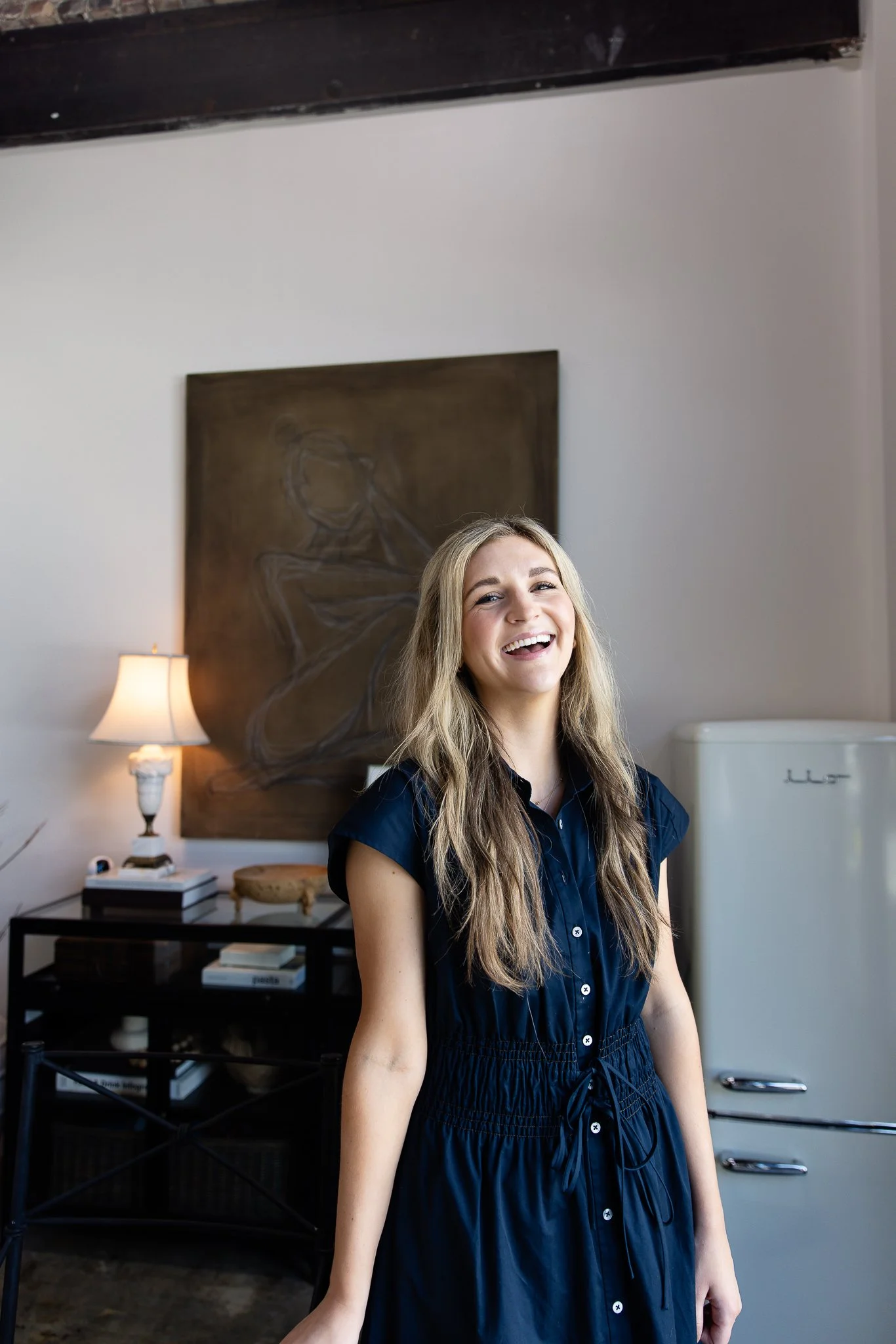 A young woman with long blonde hair smiling inside a room with a dark-colored painting and a vintage-style refrigerator.
