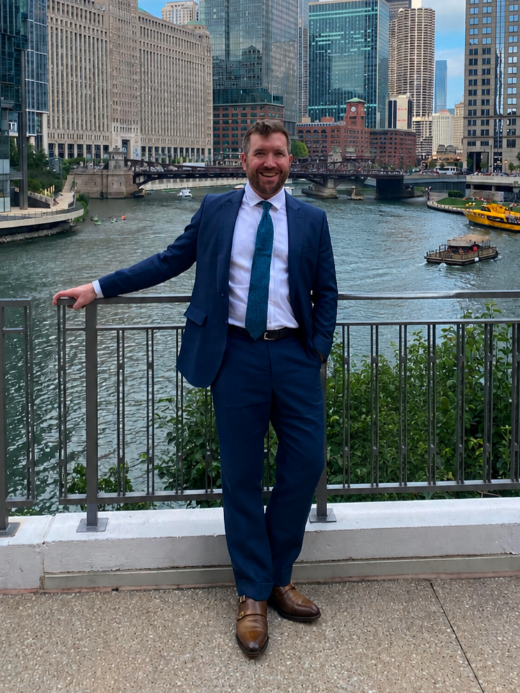 Man in a blue suit smiling, standing on a riverwalk with Chicago skyscrapers and bridges in the background.