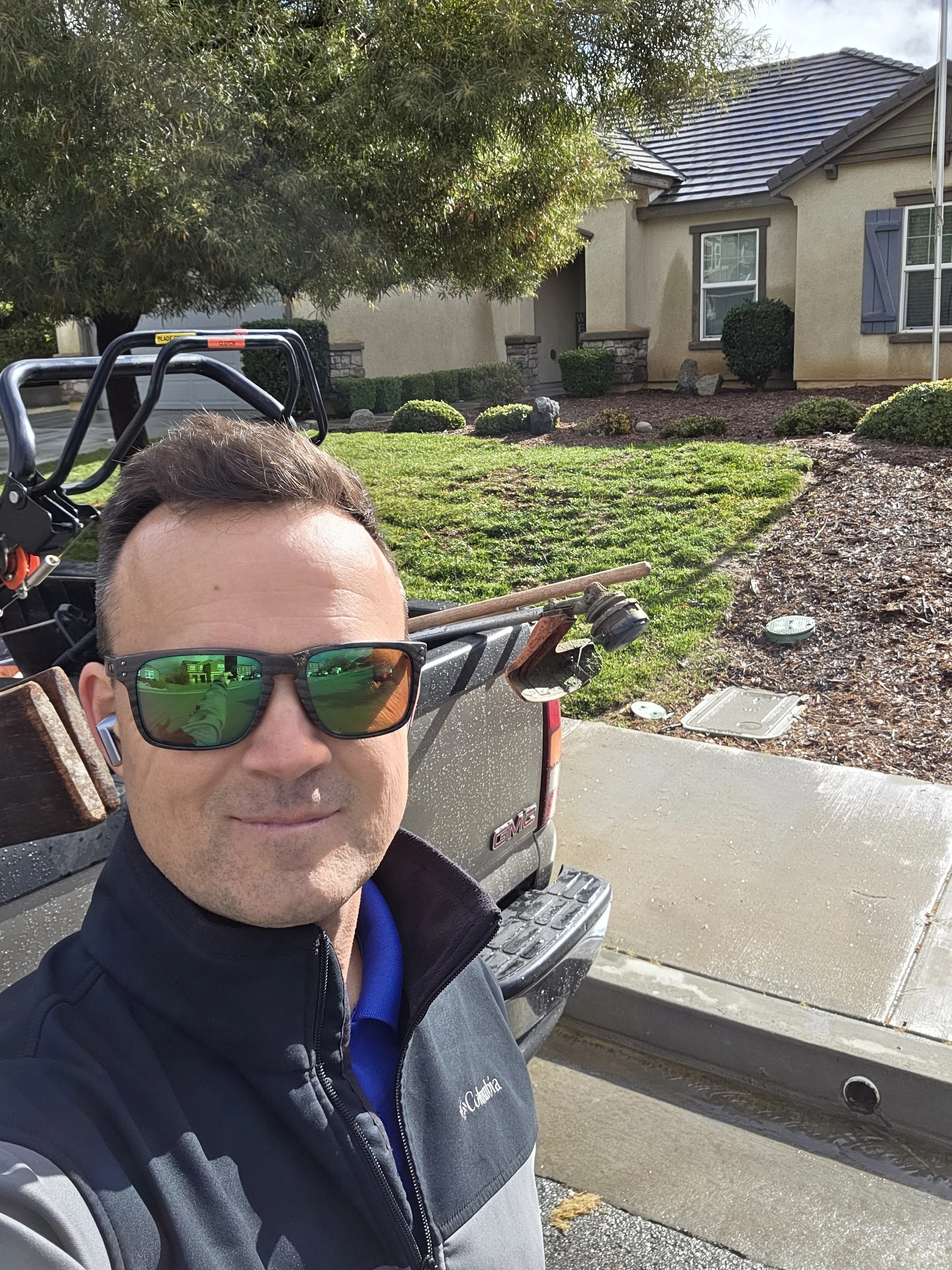 Man wearing sunglasses and jacket taking a selfie in front of a suburban house with a pickup truck nearby, a lawn, and landscaping visible.