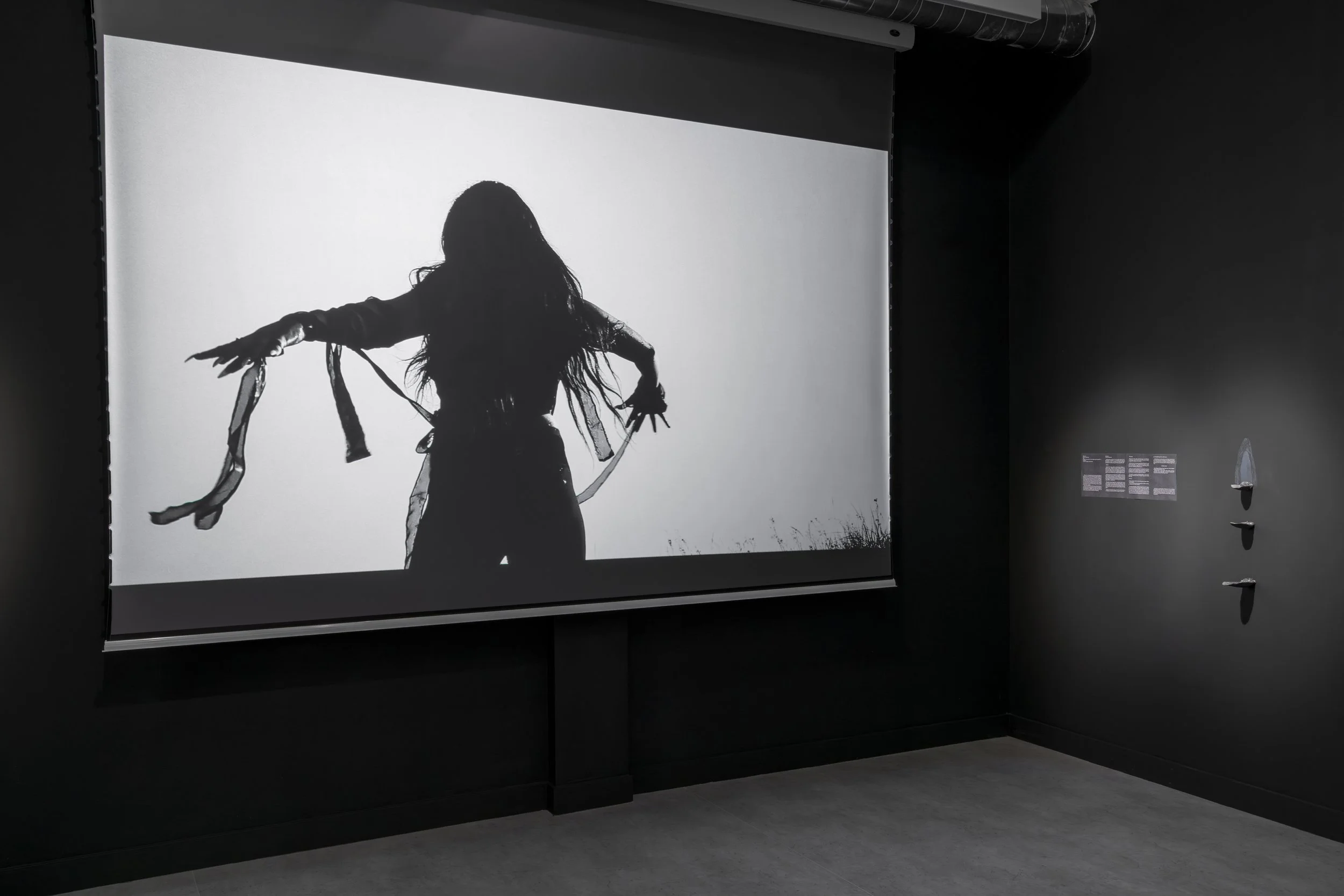  Installation view,  Vulnerable Pleasures , Gallery Gachet, Vancouver, 2025. Left: Hana Amani,  Lilith,  2022, HD video, 4 minutes. Right: Tiffany Law,  Sentimentary formation , 2024, graphite and eraser dust on carved gypsum panel suspended in a woo
