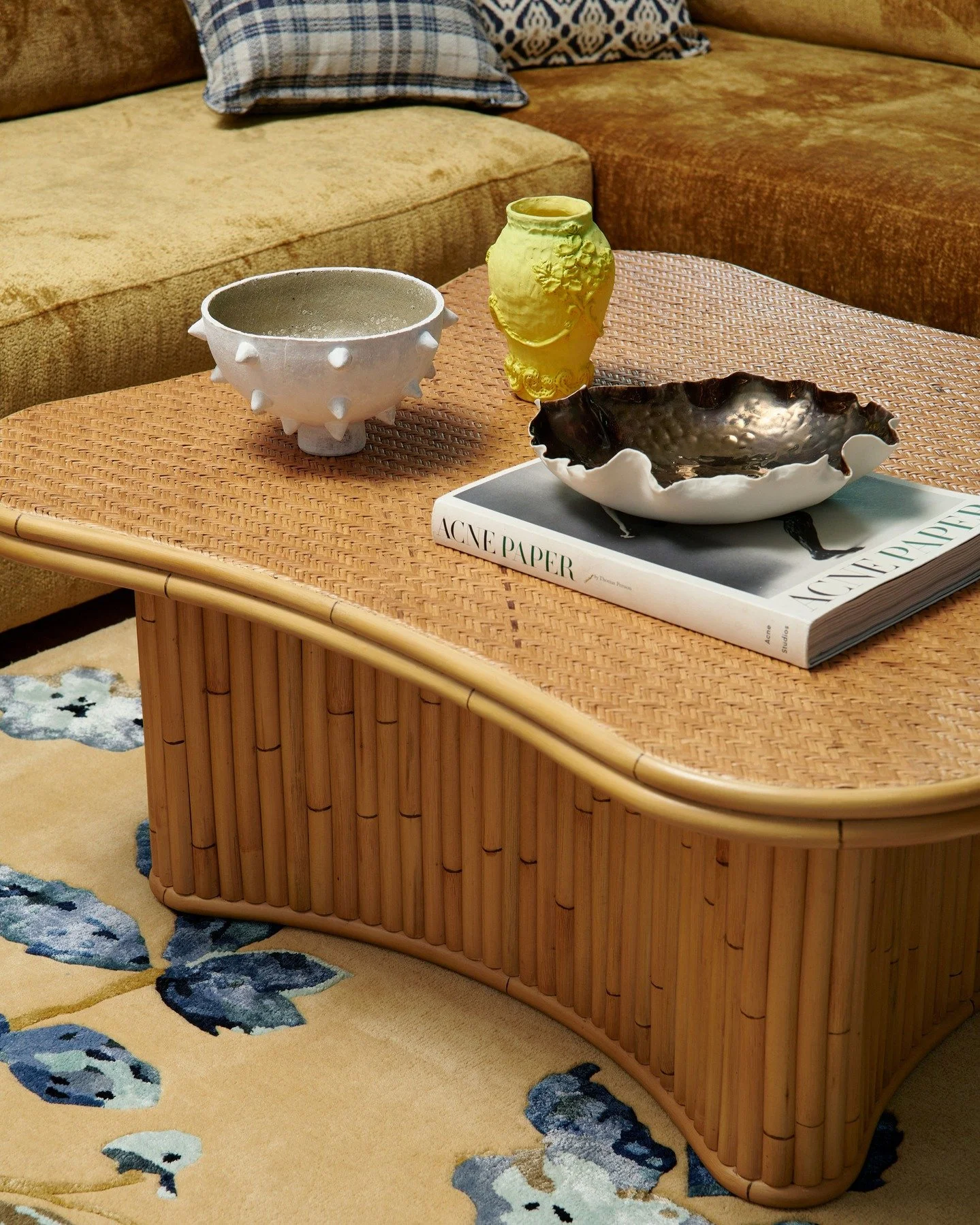KILLARA HOUSE - A woven rattan coffee table curves gently above a hand-tufted custom floral rug. Sculptural ceramics and a yellow vessel sit alongside Acne Paper, their textures set against the plush gold upholstery of the sofa. A study in material c