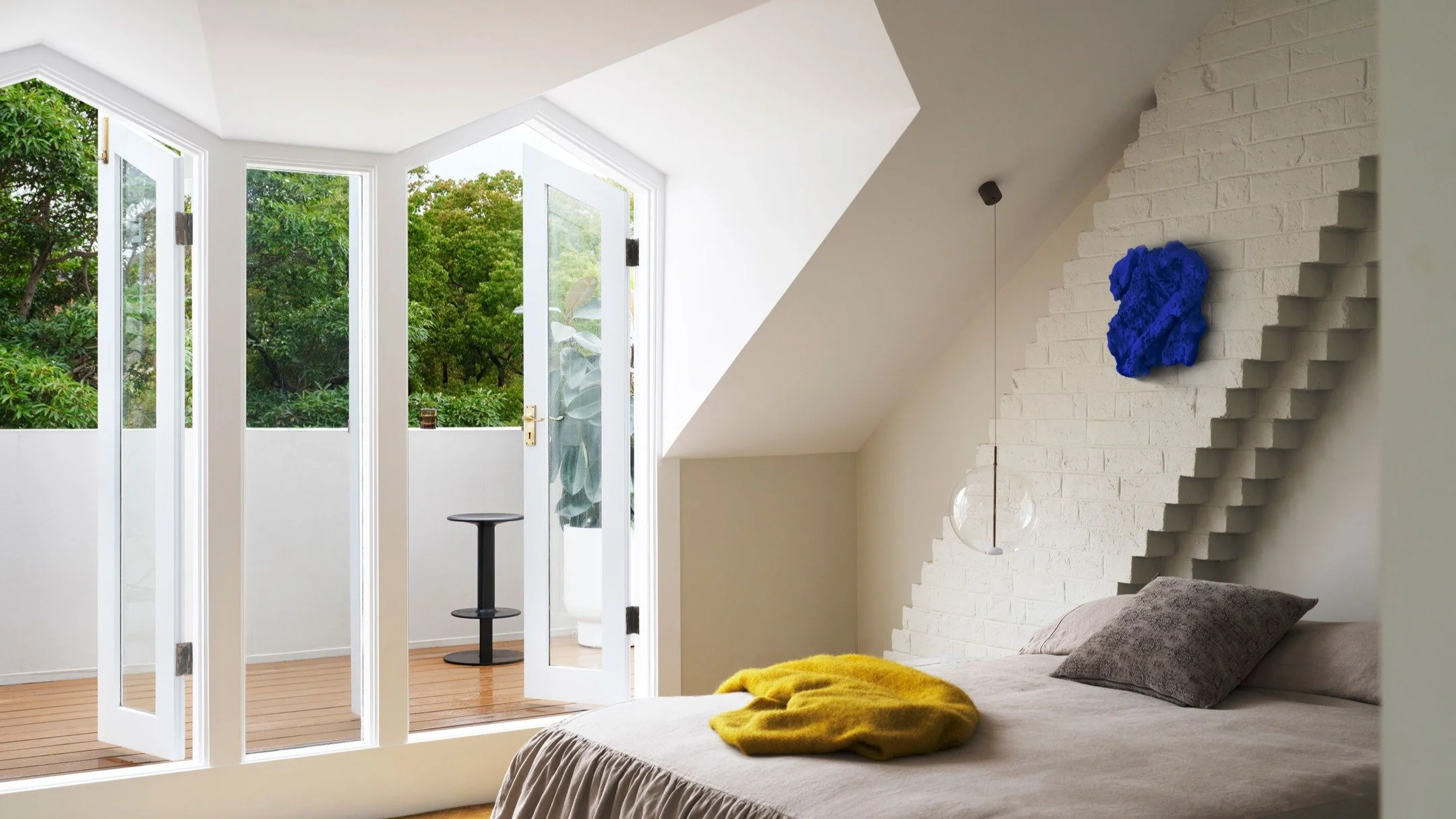 CHIPPENDALE TERRACE HOUSE &mdash; A sculpted ceiling opens to a private terrace through tall French doors, framing the treeline beyond. Inside, textured brickwork steps upward behind the bed, punctuated by a vivid cobalt artwork. Linen bedding and a 