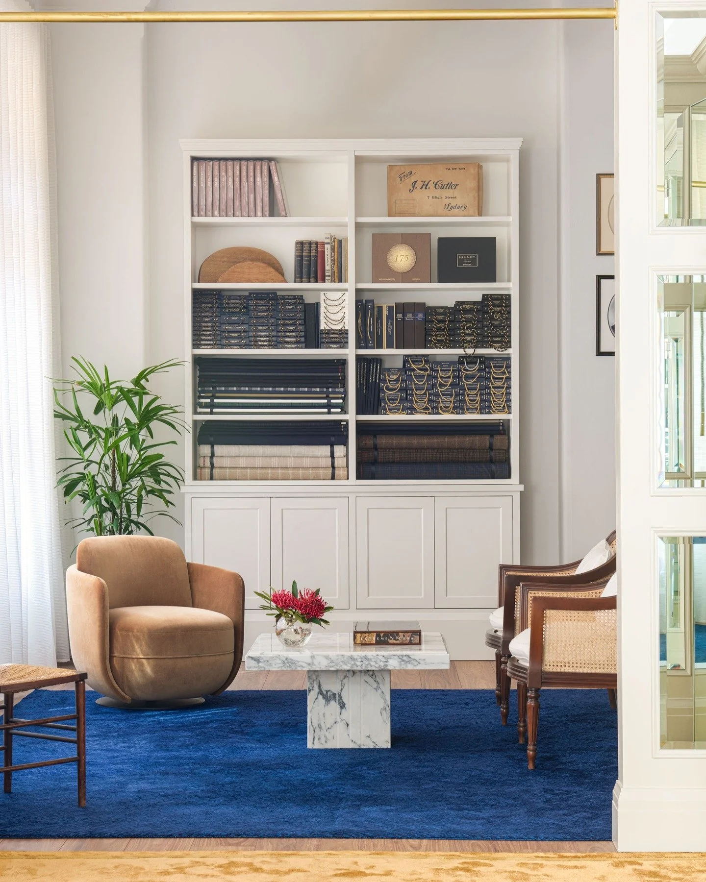 J H CUTLER - the showroom layers cobalt and gold underfoot, grounding a marble plinth table and seating in warm velvet and cane. Behind, shelves are stacked with suiting cloths and archival boxes, a backdrop that speaks to the craft and history of be