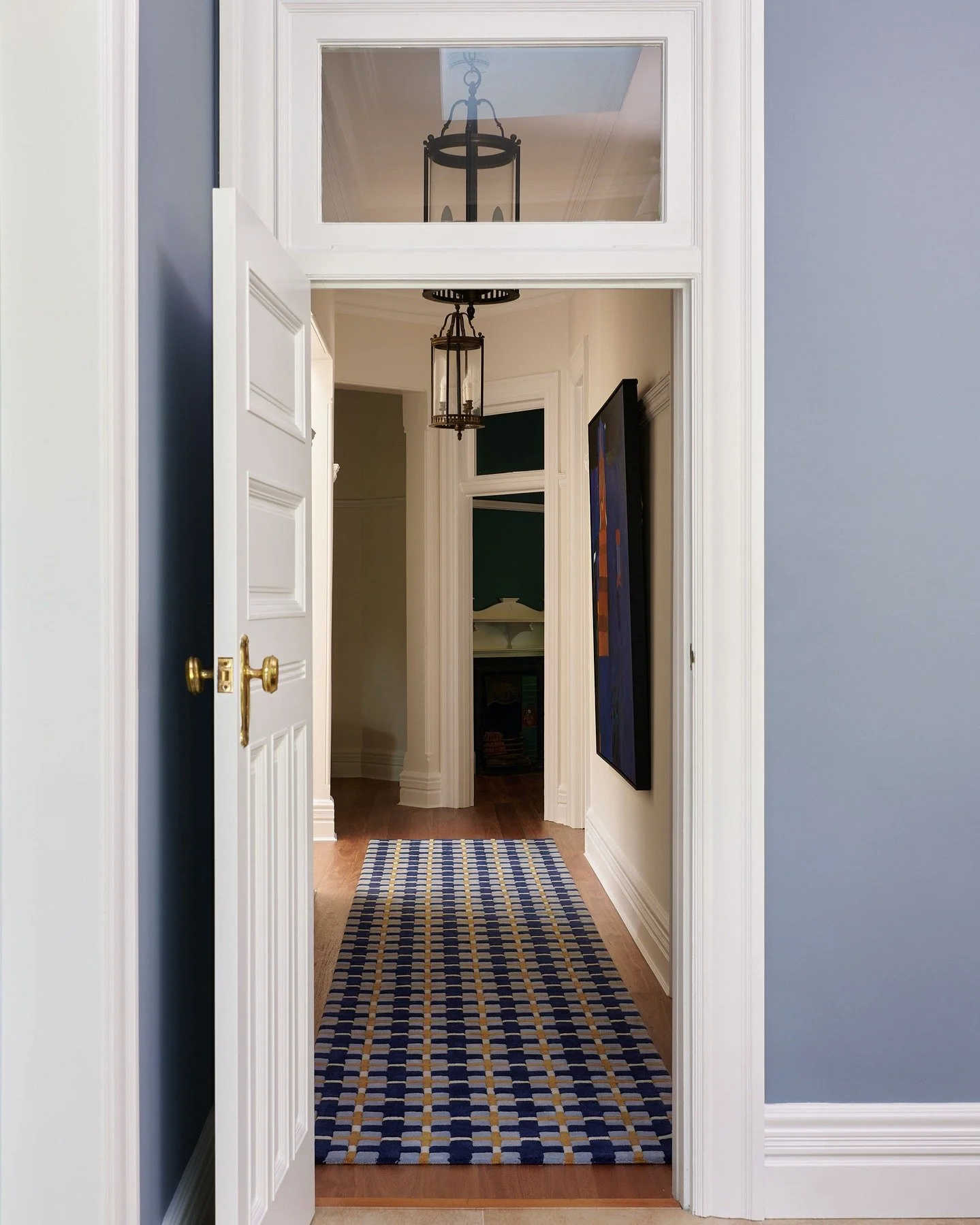 KILLARA HOUSE - A view through the long hallway at Killara House sets up a rhythm and proportion: white millwork against soft blue walls, brass door hardware catching the light. Our custom geometric runner leads you through the corridor, its cobalt a