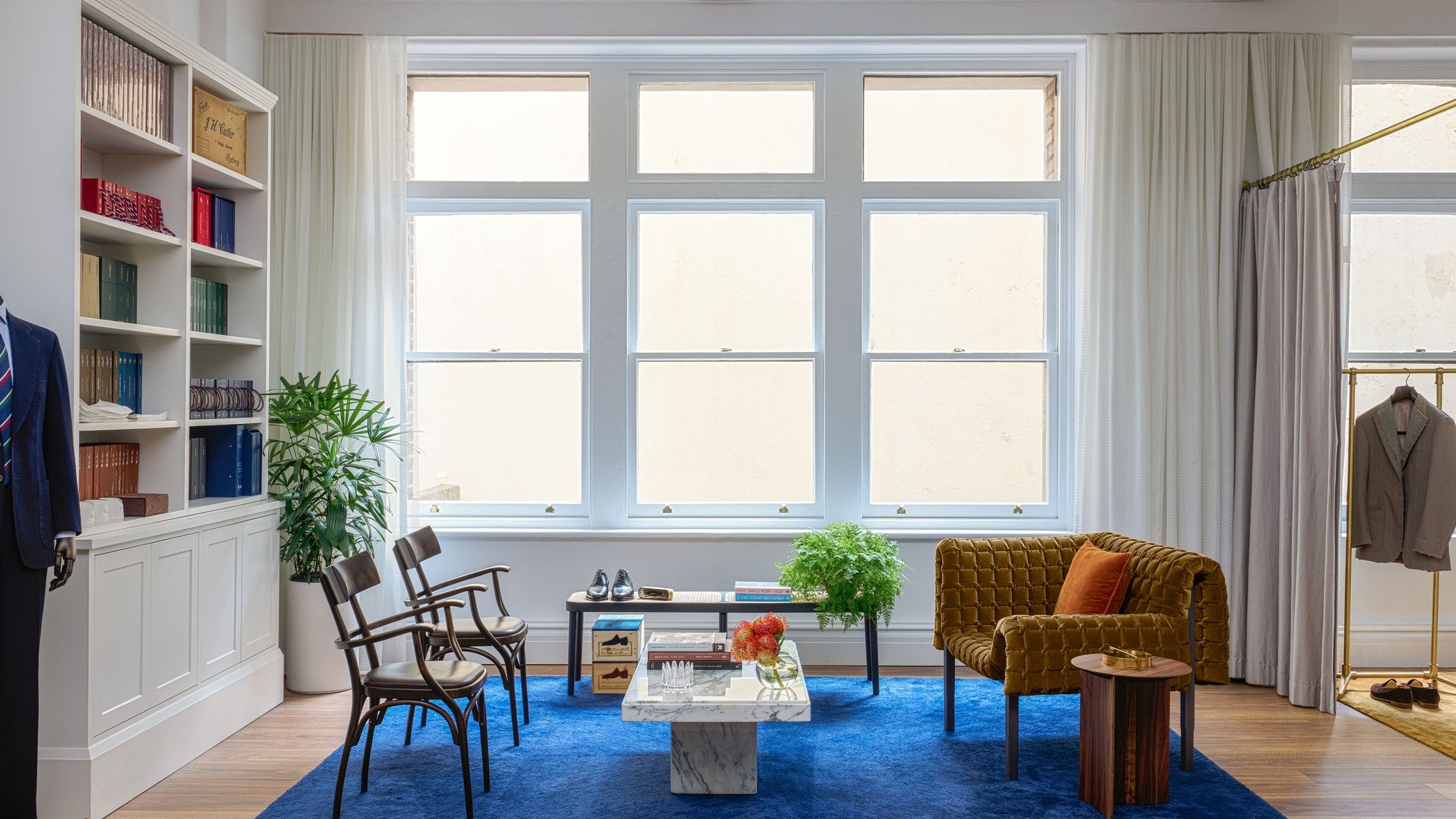 J.H. CUTLER &mdash; A fitting room arranged as a place to pause. Cobalt custom rug grounds a mix of vintage chairs, velvet seating and marble table, set against tall sash windows and tailored joinery. A room designed around conversation and craft.

I