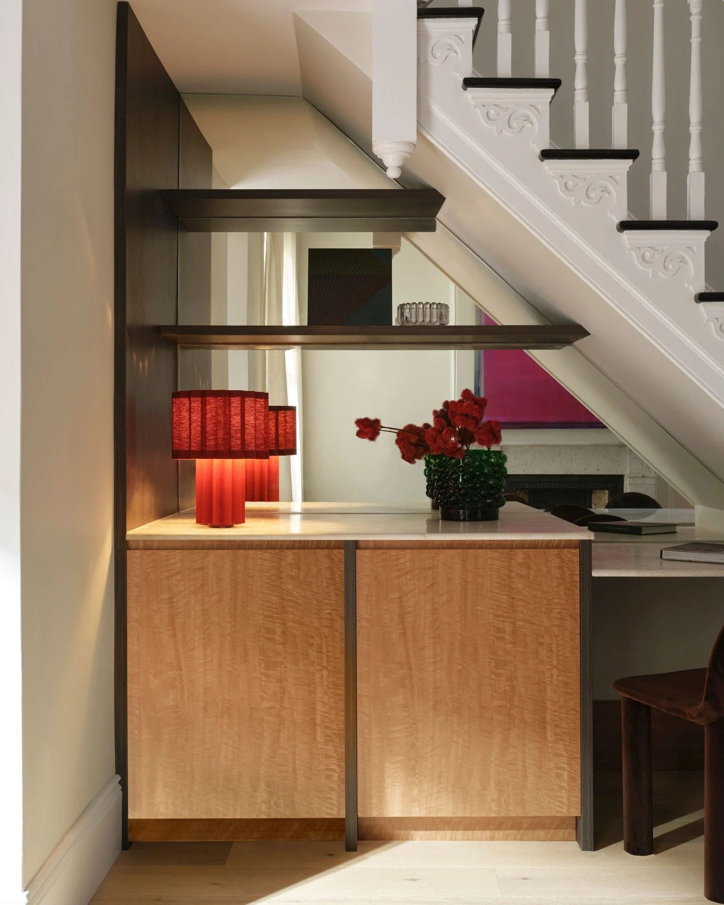 CHIPPENDALE TERRACE HOUSE &mdash; A compact bar built into the void beneath the stair. Figured timber doors sit within a darker frame, topped with stone and softened by open shelving. A red pleated lamp shifts the palette, turning this corner into a 
