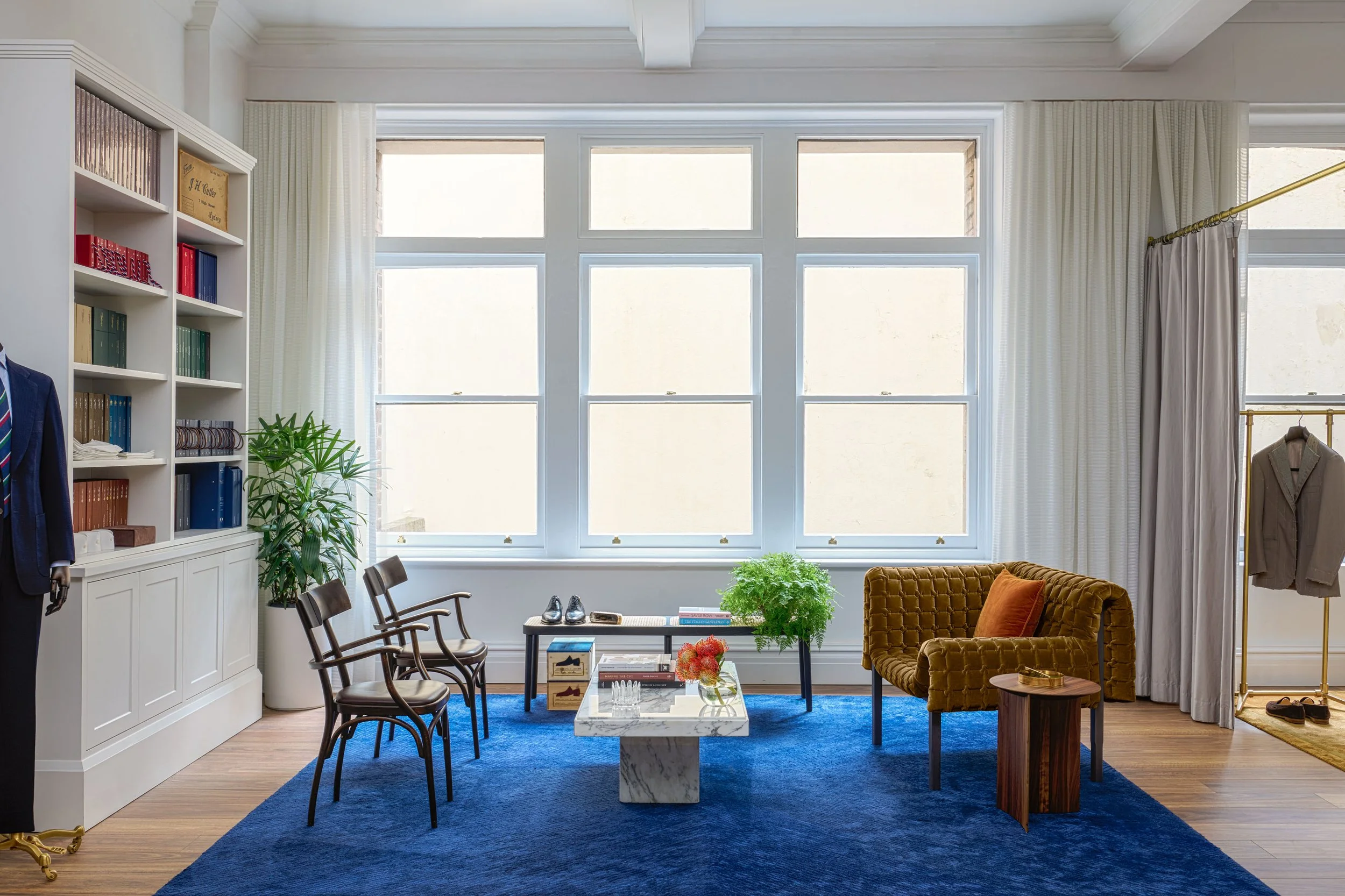 Lounge seating area with mustard velvet chair, blue carpet, and bespoke tailoring accessories at J.H. Cutler atelier in Sydney.