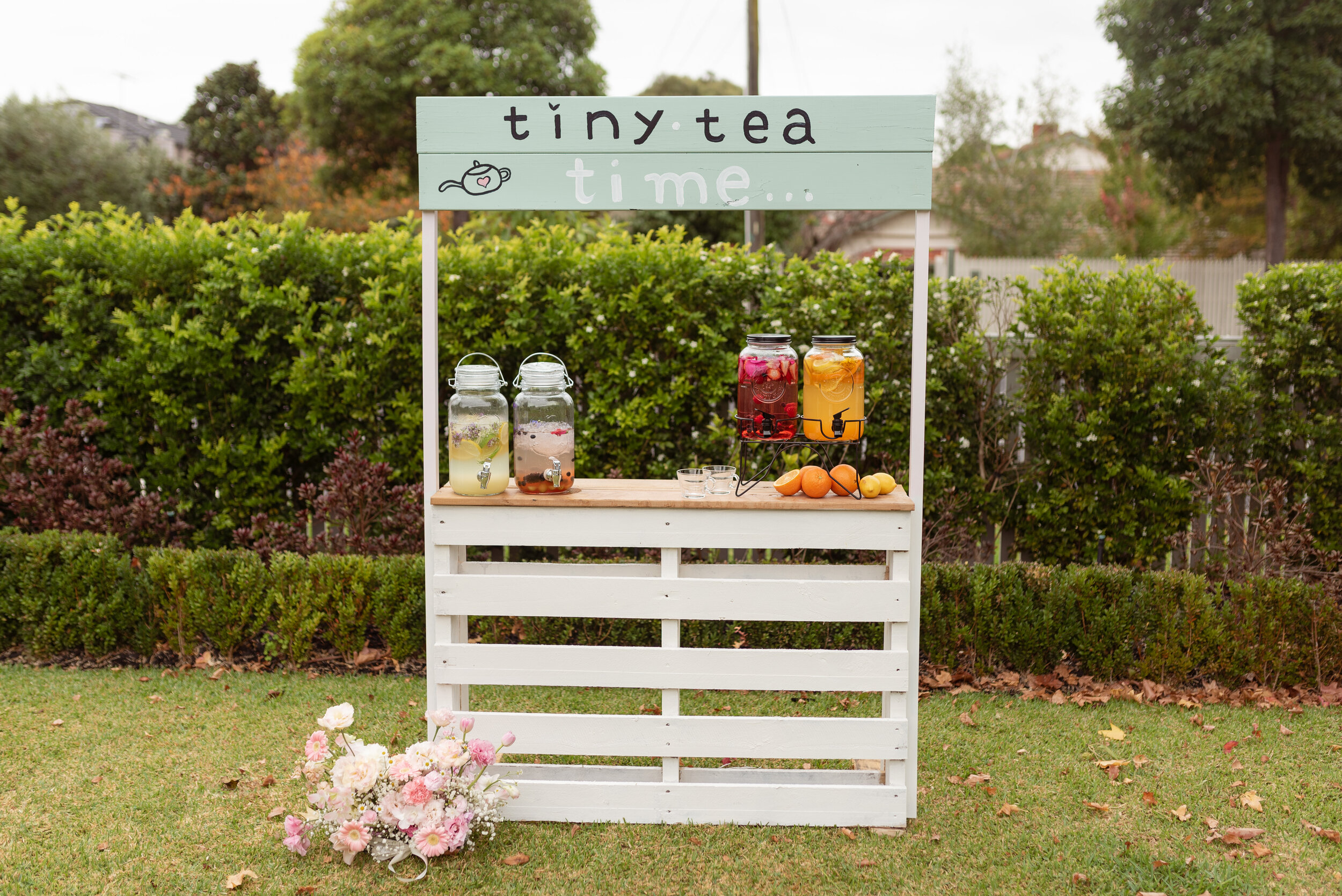 A drinks stand in a garden, sittong on green grass with pink and white florals at the foot of the stand. The sign above the stand reads, "Tiny tea time...". Drinks dispensers full with fruity, colourful tea, with fresh oranges next to them on the table. We were thrilled to be able to provide creative direction for the entire process of the brand shoot, and we worked closely with a number of eco-friendly suppliers to come together and create a delightful shoot for Tiny Tea Kids.
