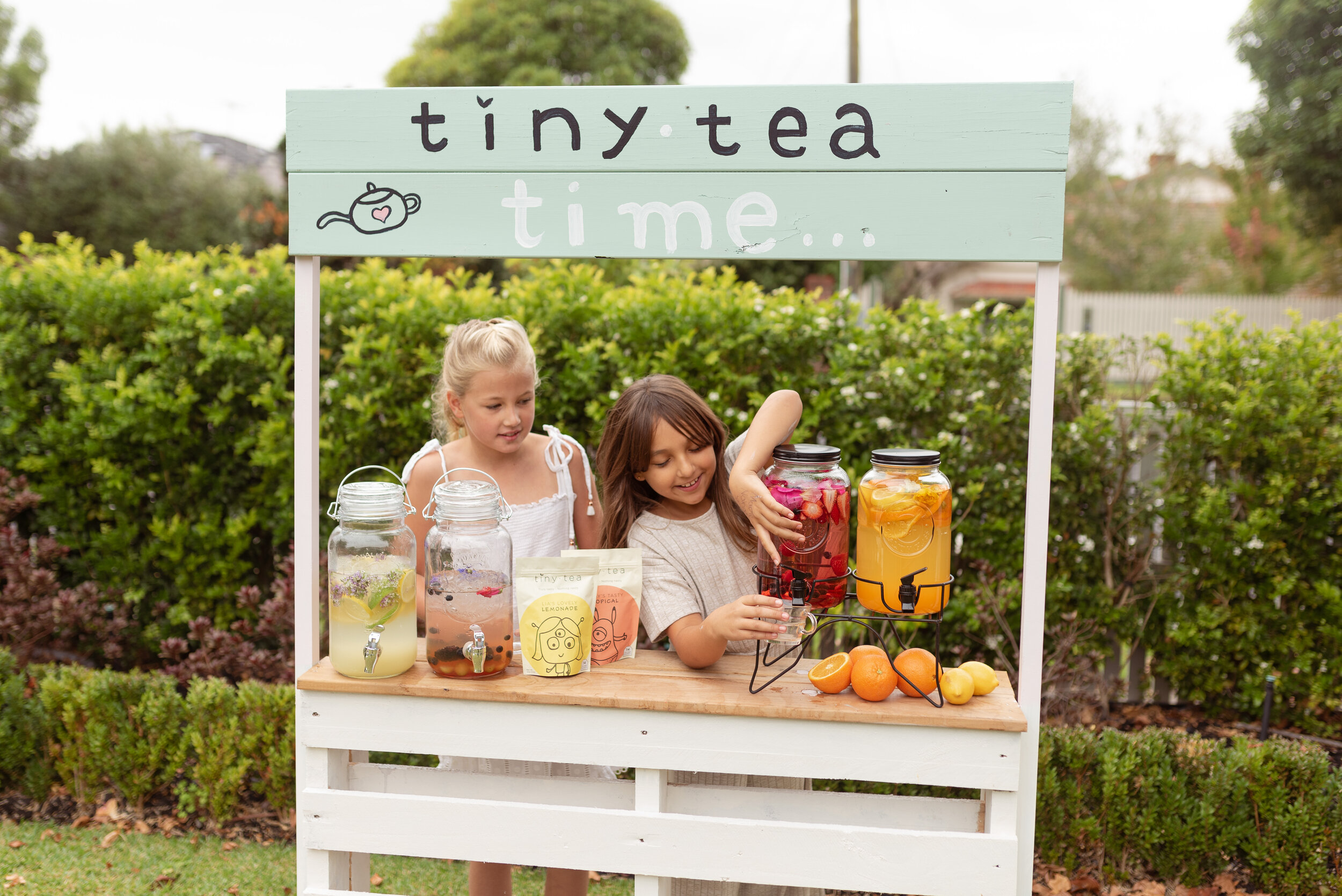 Two children in the tea stand pouring tea from the dispenser. Tiny Tea pouches sit on the table alongside fresh fruit. 
The children loved our custom homemade drink stands and really got stuck in with the cold brewed tea fun!