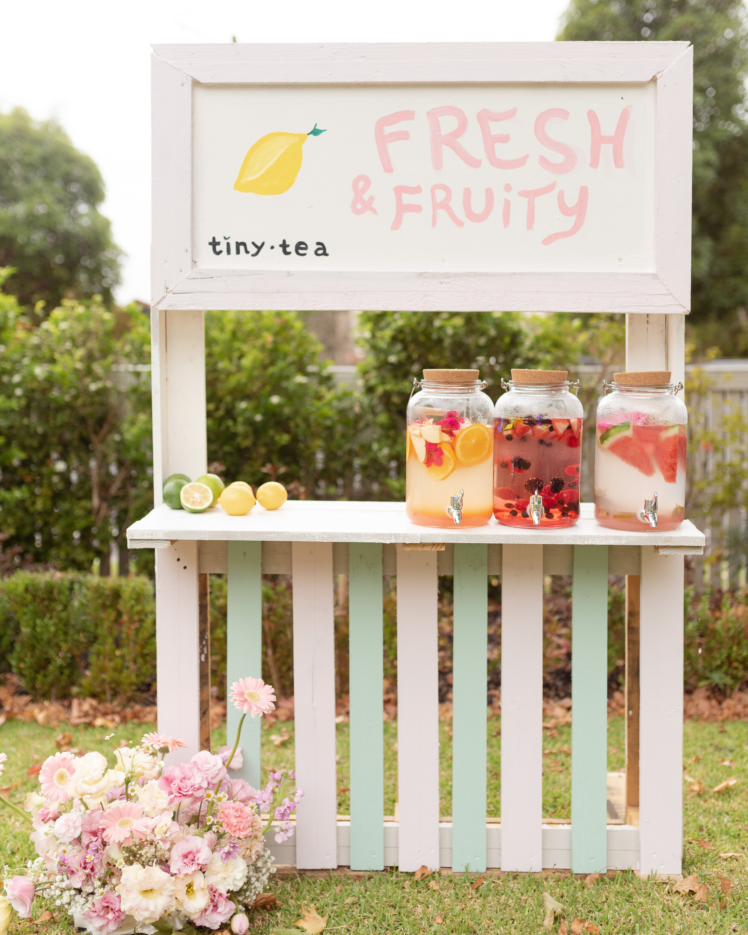 Another drinks stands, this one reading, "Fresh and Fruity, tiny tea". With fresh cut lemons and lime, next to full drinks dispensers, full with tea and fresh fruit. 
Starting the day with an outdoor garden party, we worked with the brief in mind - that being modern, fun and fresh, with light pastel colours - to create a stunning picnic setting.Complete with carefully curated florals, decals, dinnerware, stationary and tasty treats, chalk and colouring-in stations, a tea station and tea stands - of course - and the most dreamy cubby house that we definitely all wanted in our own backyard, it really was the perfect party for the kids!