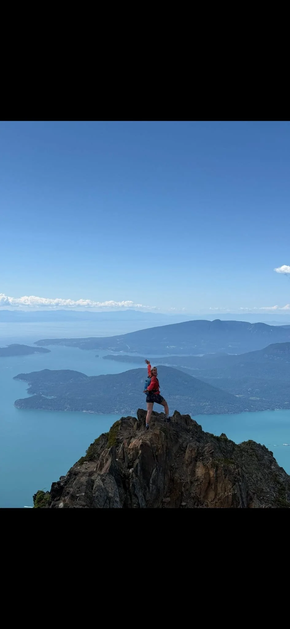 Main Street RMT Marina Baker standing at the top of Brunswick mountain on a clear day, with islands in the background and a blue sky. RMT Marina is wearing a red jacket. RMT Marina is carrying a blue backpack.