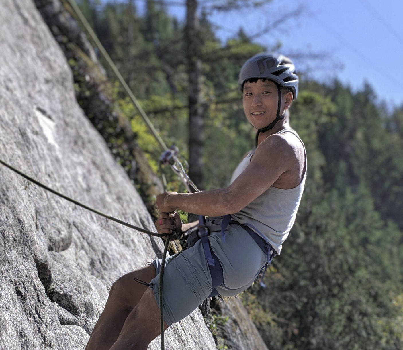 Main Street RMT Robert Young climbing, wearing a harness and a helmet. RMT Robert is also wearing a white tank top and green shorts. THere are trees and blue sky in the background, while RMT Robert is hanging on a rope on belay.