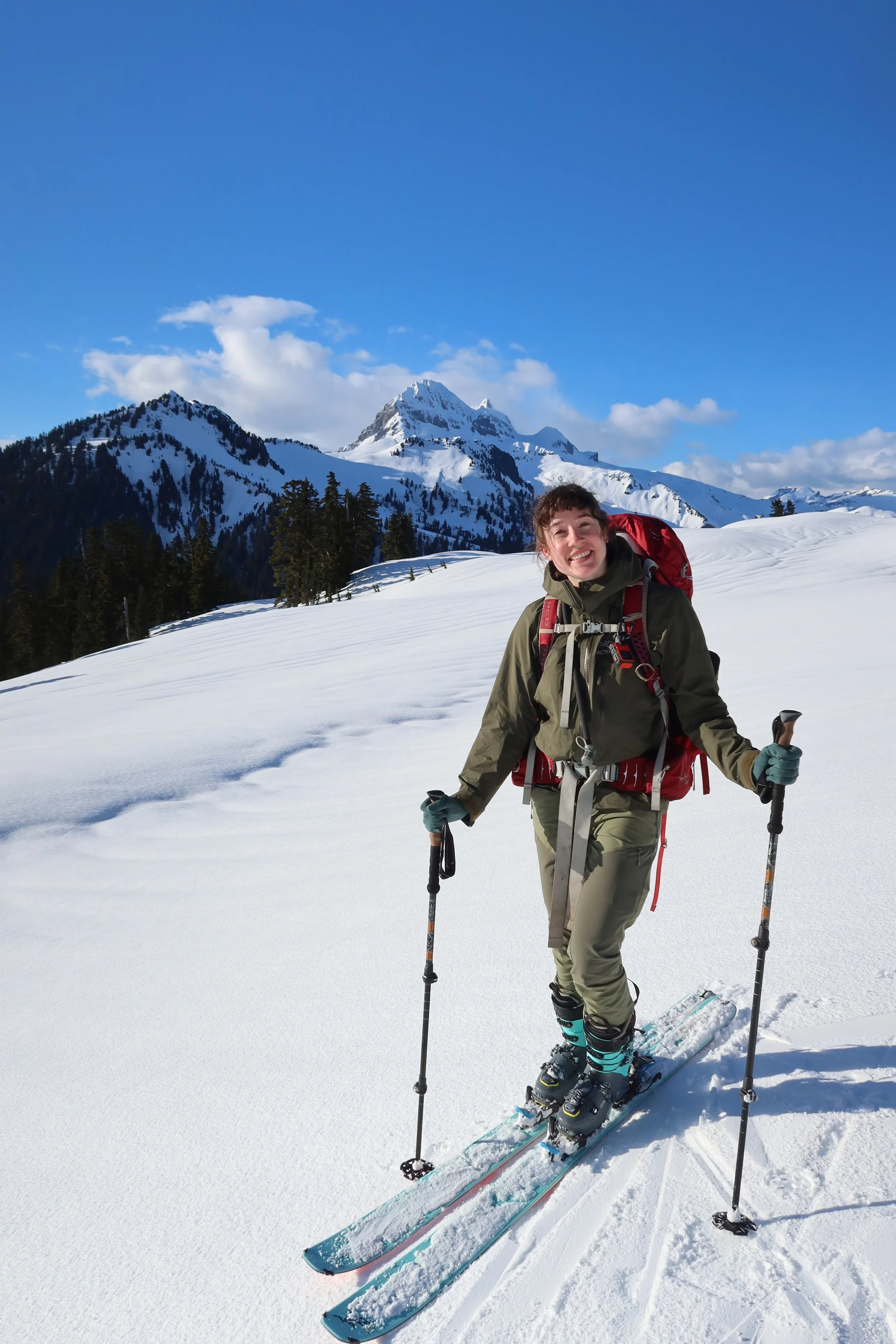Physiotherapist Sophie Provenzano backcountry skiing with blue sky and mountains in the background. Physiotherapist Sophie is wearing a green jacket, green pants, and carrying ski poles and a red backpack.