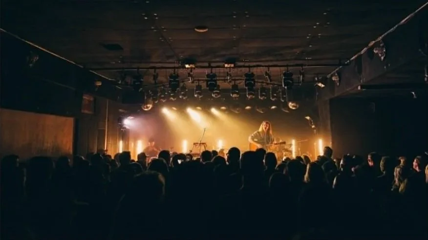 A live music performance on stage with Benjamin William Hastings and cellist Caleb Yang, audience watching in a dimly lit venue with stage lighting.