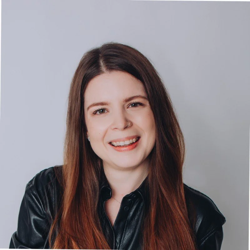 A woman with long brown hair, wearing a black leather jacket, smiling and making a playful face against a plain gray background.
