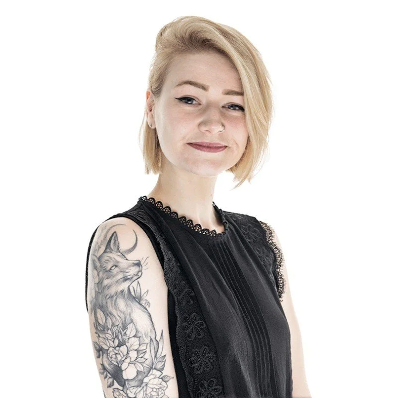 A woman with blond hair, wearing a black sleeveless top with lace details, showing a large tattoo of a fox and flowers on her left arm, and smiling at the camera.