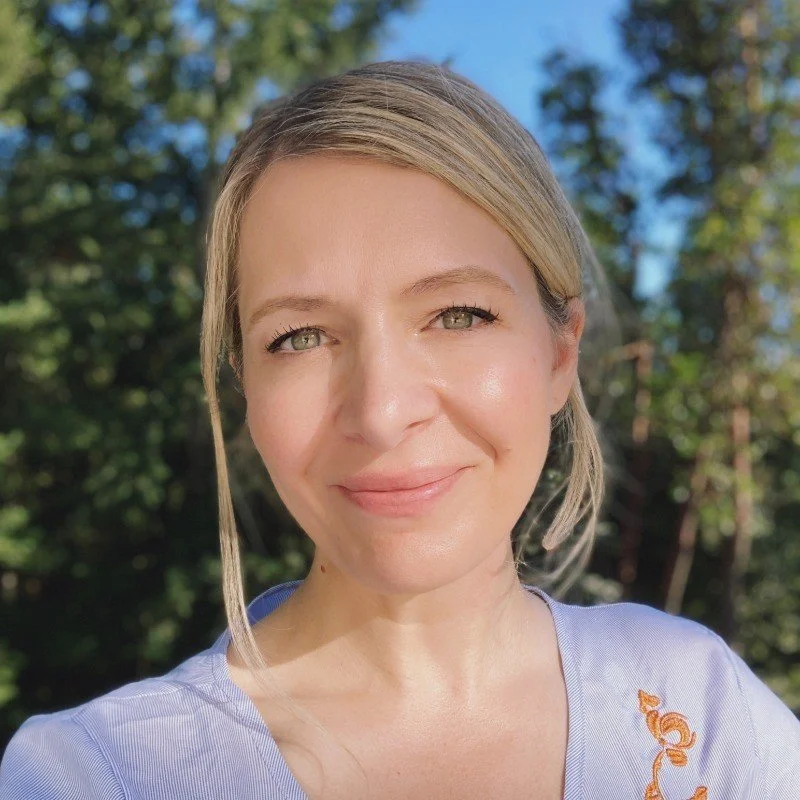 A woman smiling outdoors with green trees in the background.