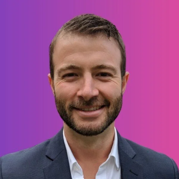 Smiling man with a beard and short brown hair, wearing a dark suit jacket and a white dress shirt, standing against a pink and purple gradient background.