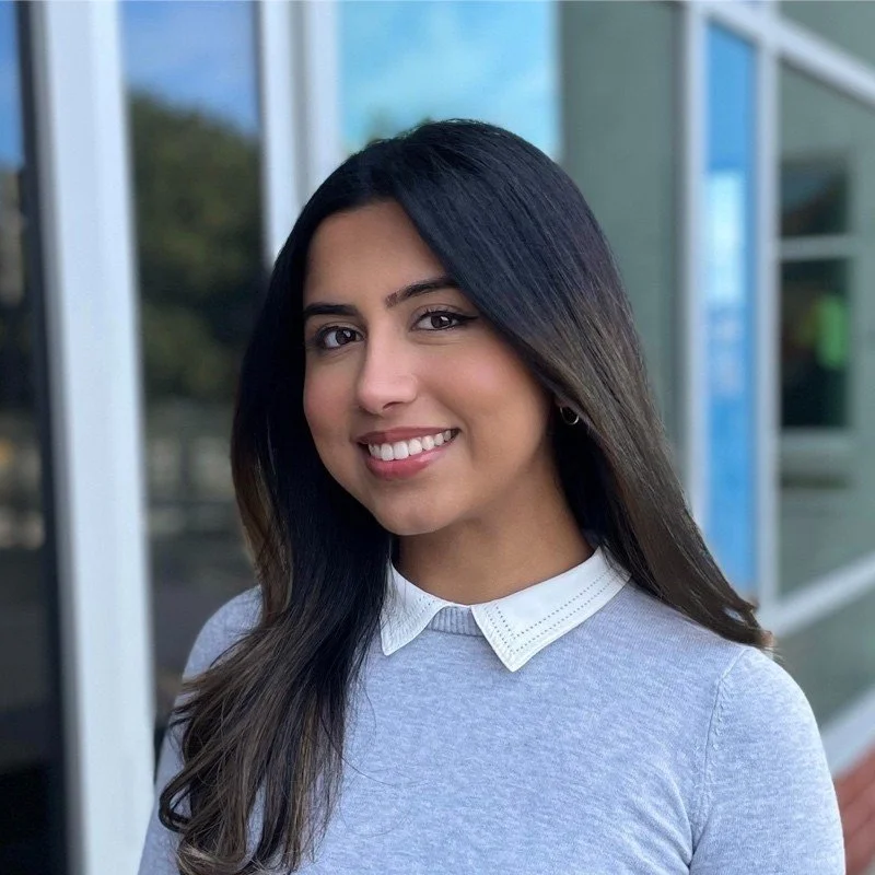 A young woman with long dark hair, smiling, standing outdoors near glass windows, wearing a light gray top and white collared shirt.