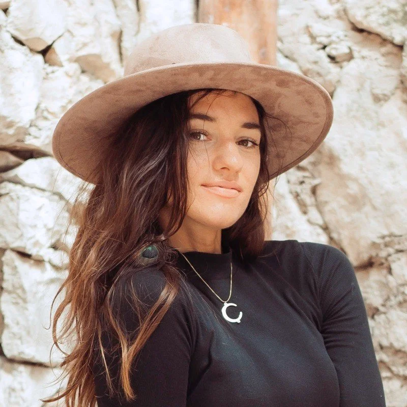 A woman with long brown hair wearing a wide-brimmed beige hat, black shirt, and a necklace with a letter 'C', standing against a rocky background.