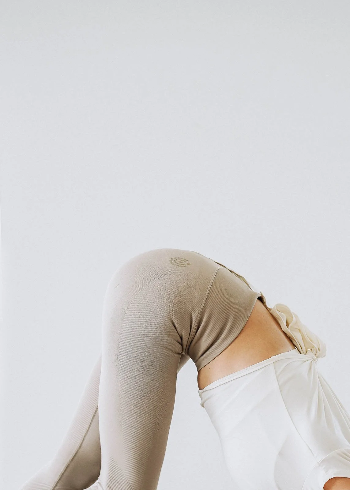 Person beim Yoga in neutraler Kleidung in einer Haltung, Fokus auf den Rücken und Beine, weißer Hintergrund.