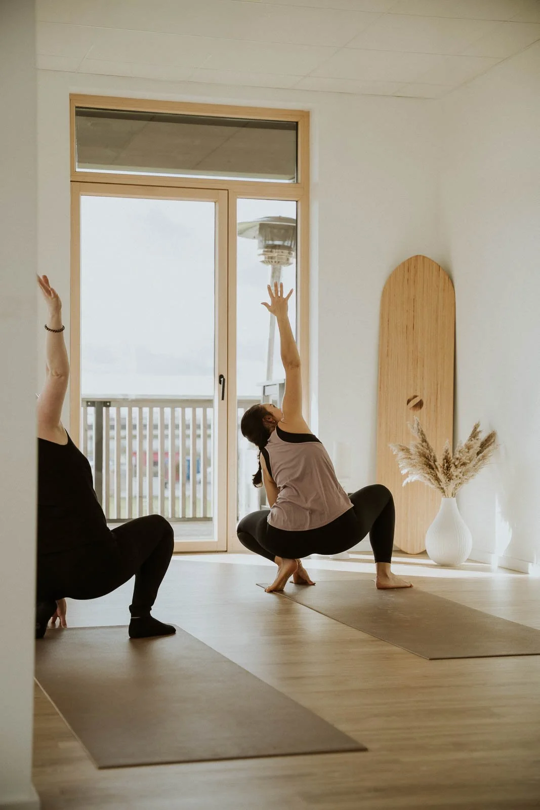Zwei Frauen machen Yoga im Wohnzimmer. Das Foto ist in warmem Licht aufgenommen, neben einem großen Fenster steht eine Vase mit Trockenblumen.