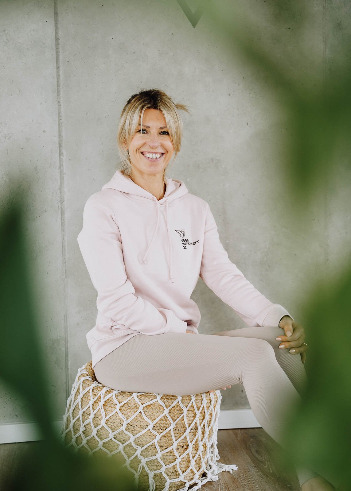 Eine lächelnde blonde Frau in einem hellrosa Hoodie sitzt auf einem Korb gegen eine Betonwand. Sie ist entspannt und schaut in die Kamera.