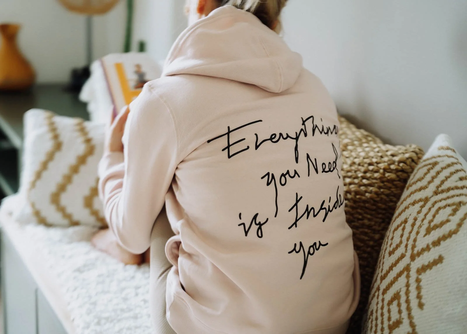Person in hellbeigem Hoodie mit schwarzem Aufdruck, der sagt "EveryTime you Need it Inside You" auf dem Rücken, sitzt auf einem Sofa mit weißen Kissen mit goldfarbenem Muster.