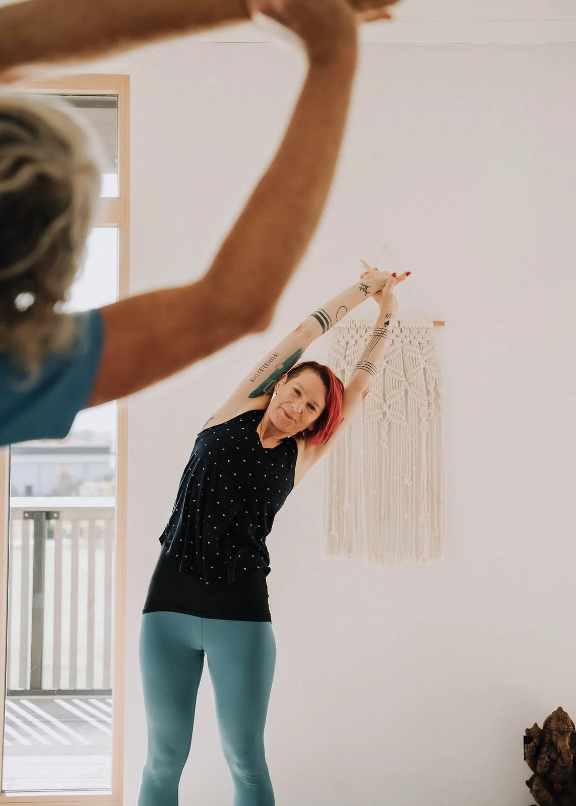 Frau beim Yoga dehnt sich in einem hellen Raum mit Wandbehang. Sie trägt eine schwarze Top und blaue Leggings, hat rote Haare und Tattoos auf den Armen.