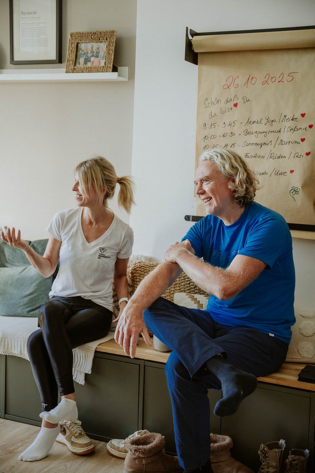Zwei Menschen, eine junge Frau und ein älterer Mann, sitzen auf einem Sofabank in einem Raum. Sie lachen und unterhalten sich. Hinter ihnen ist eine Pinnwand mit einem Terminplaner und einer Notiz vom 26.10.2025.
