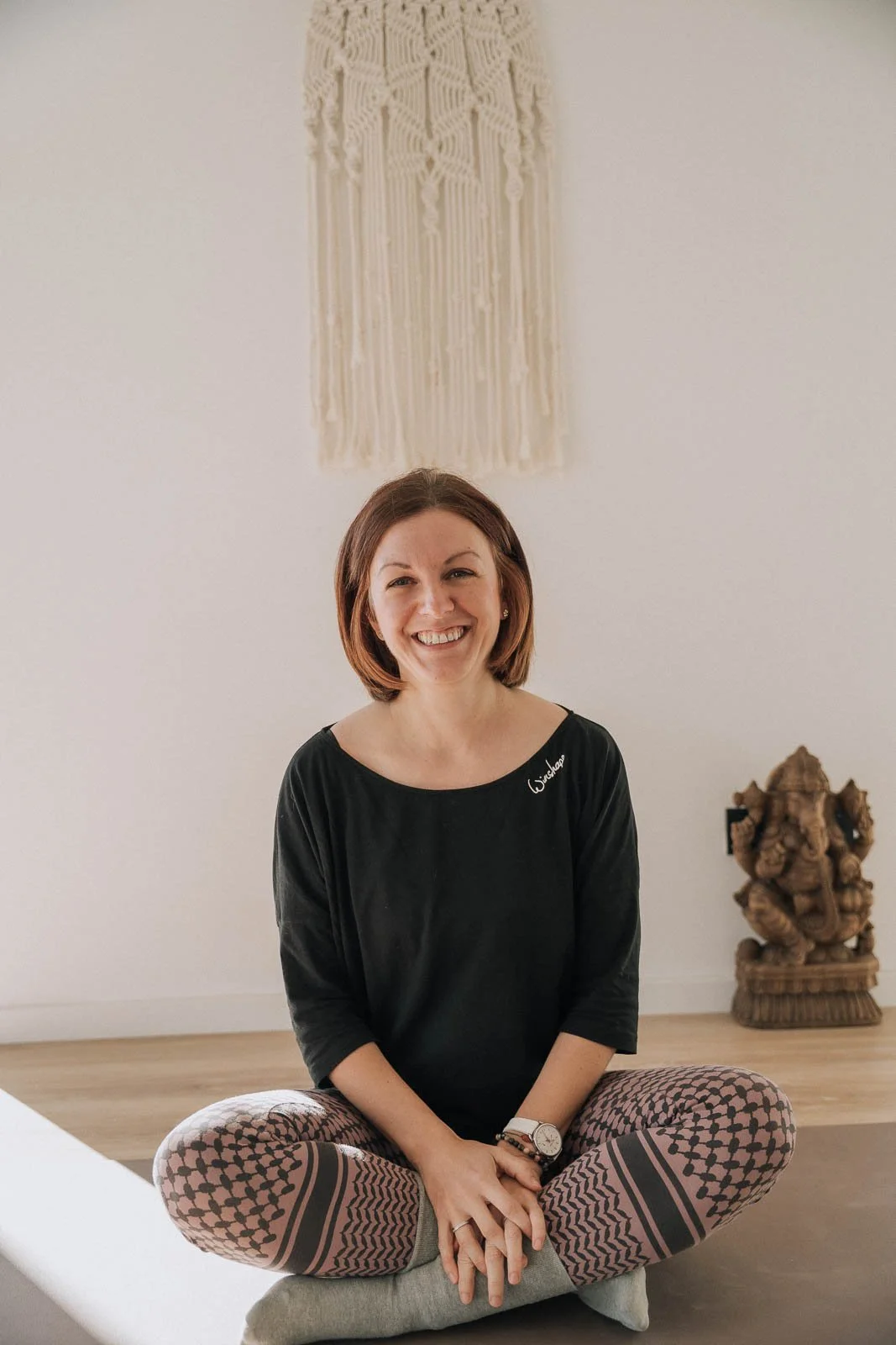 Frau sitzt im Schneidersitz auf einer Yogamatte in einem Raum mit Holzboden, hinter ihr hängt eine Makramee-Wanddekoration, neben ihr steht eine geschnitzte Ganesha-Statue.