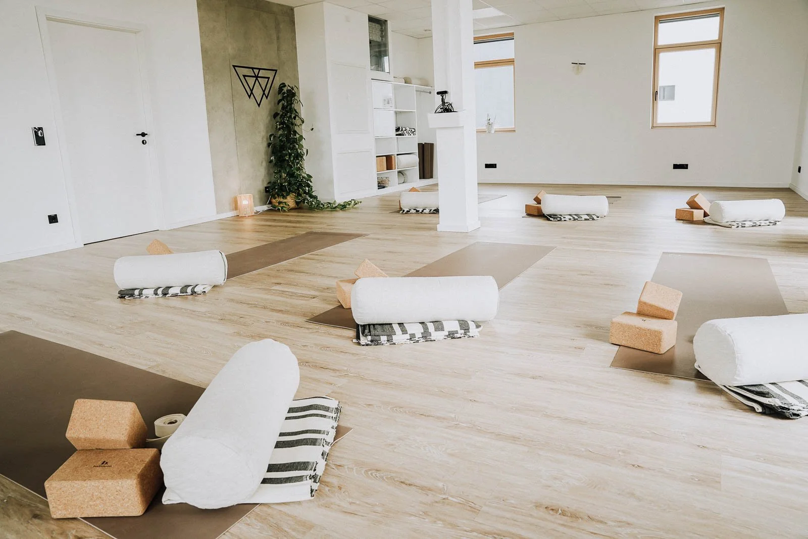 Ein heller Yogaraum mit Yoga-Matten, Yogabolstern, Decken und Ziegelsteinen auf einer Holzbodenfläche, bereit für eine Yogastunde.