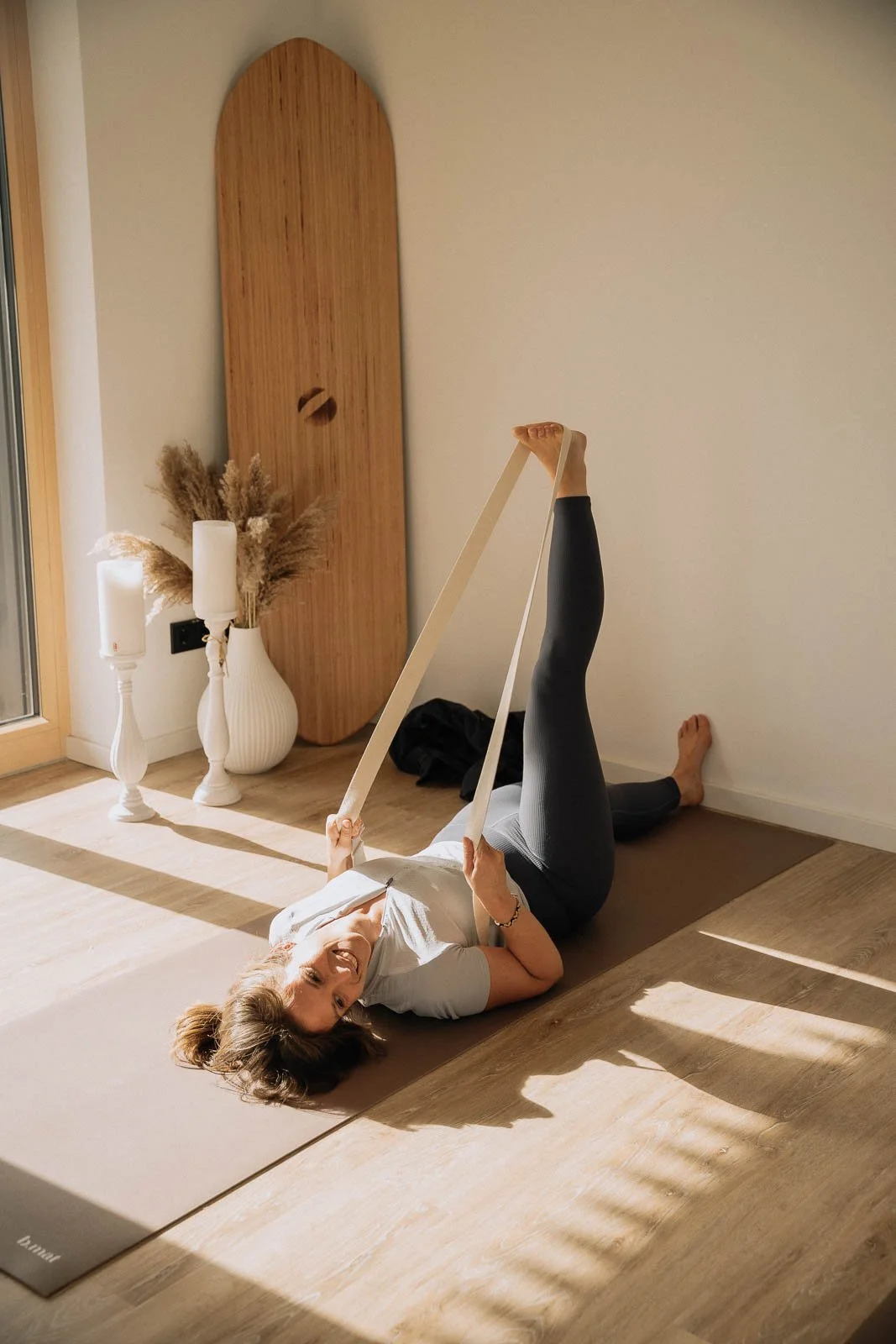 Frau macht Yoga auf Matte, hält ein Yoga-Prop in den Händen, Sonnenlicht fällt ins Zimmer