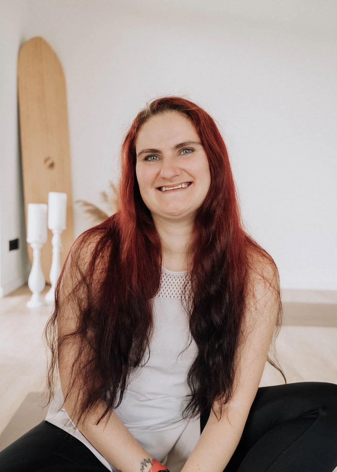Junge Frau mit roten Haaren sitzt auf dem Boden, lächelt und schaut in die Kamera, im Hintergrund weiße Wände mit dekorativen Kerzen auf einem Tisch.