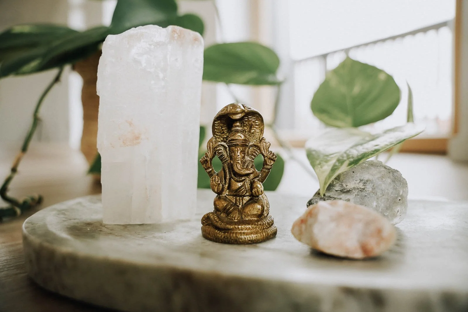 Eine Zinnfigur des Gottes Ganesha, umgeben von einer weißen Salzkristallstange, einem grünen Blatt und einem kleinen Stein auf einer runden Steintafel im natürlichen Licht.