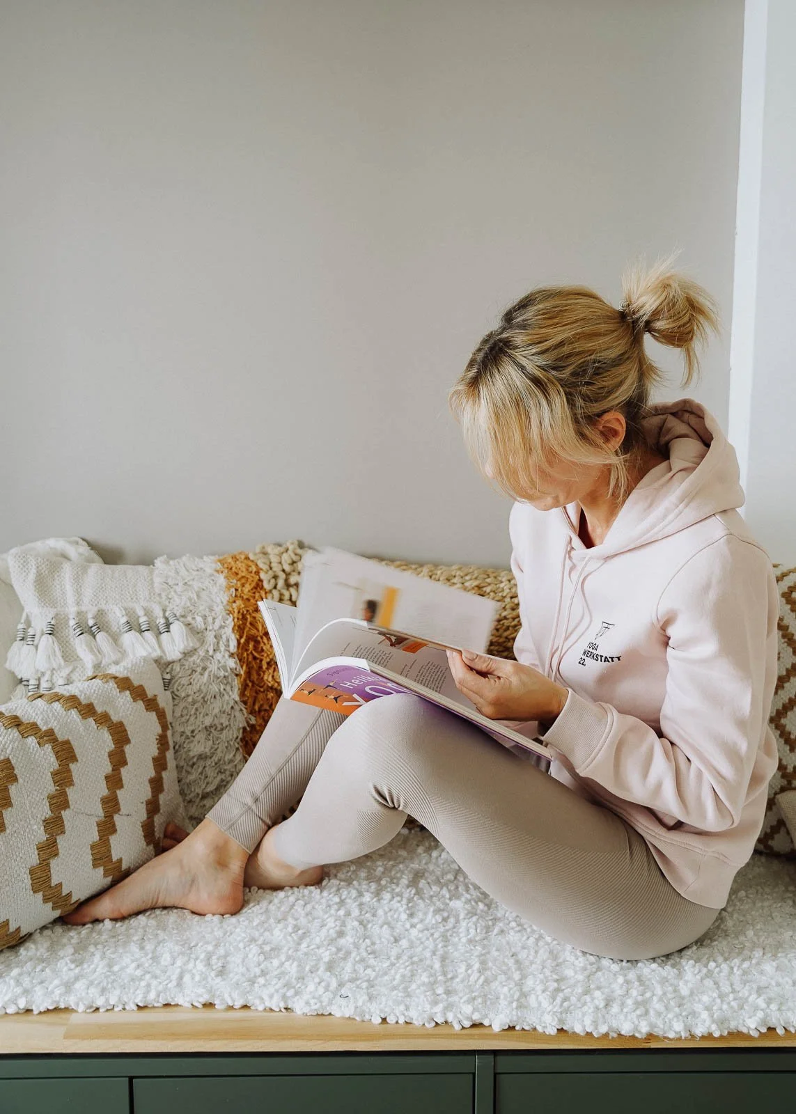Frau liest ein Magazin auf einem gepolsterten Sitz mit Kissen, trägt einen hellrosa Hoodie und Leggings, sitzt mit gekreuzten Beinen auf einer weichen Decke, vor einer weißen Wand.