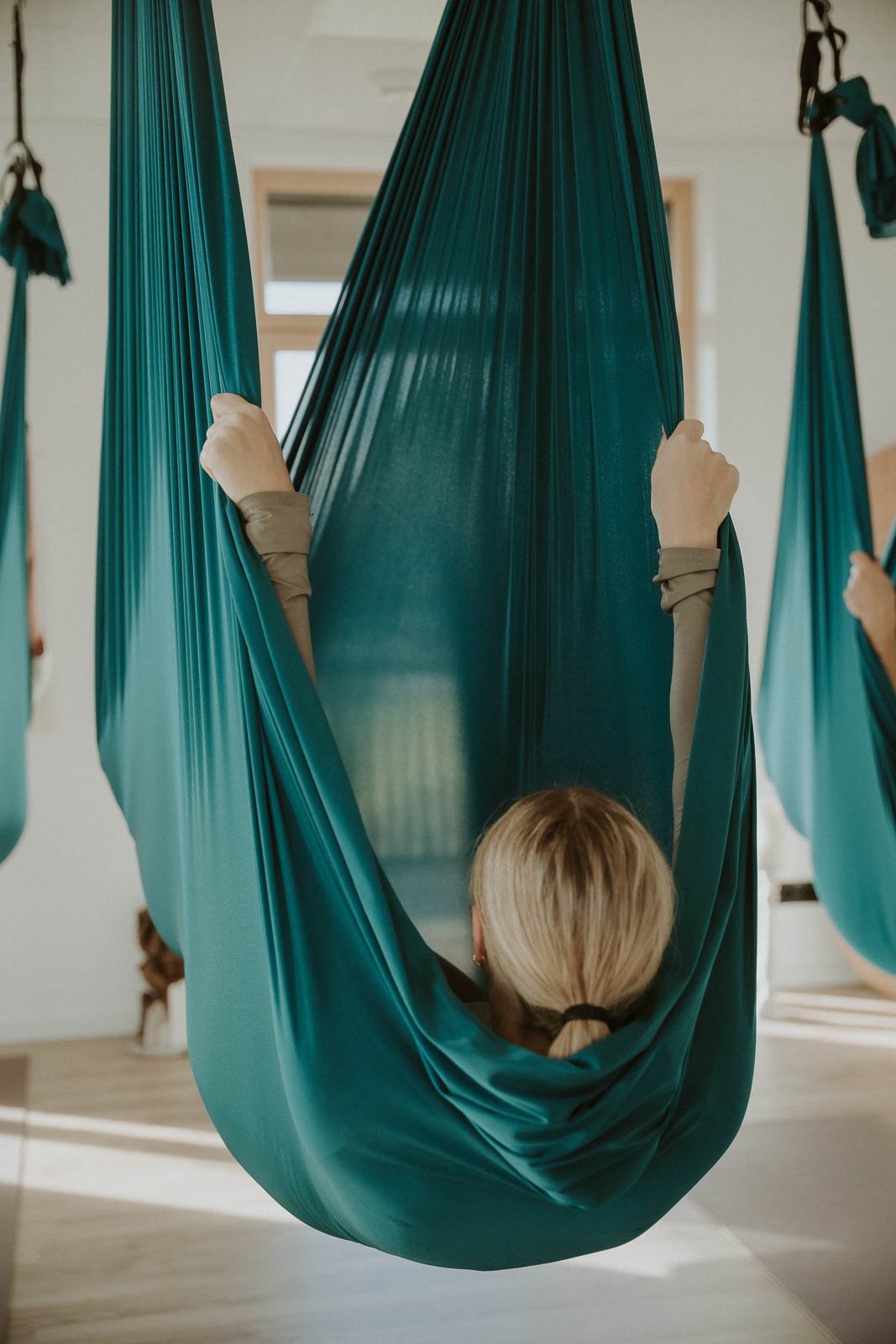 Person in einem Hängesitz aus blauer Stoff im Innenraum, umgeben von weiteren blauen Hängesitzen, mit Blick auf die Fenster im Hintergrund.