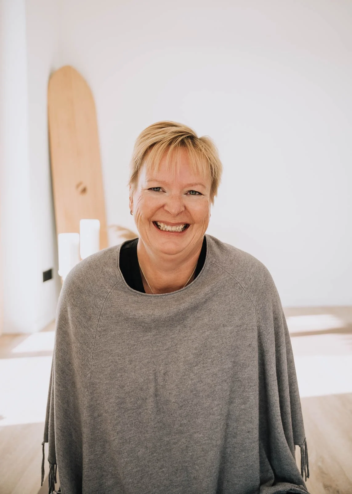 Lächelnde Person in grauem Poncho, sitzend in einem hellen Raum mit dezenten Holz- und Kerzenelementen.