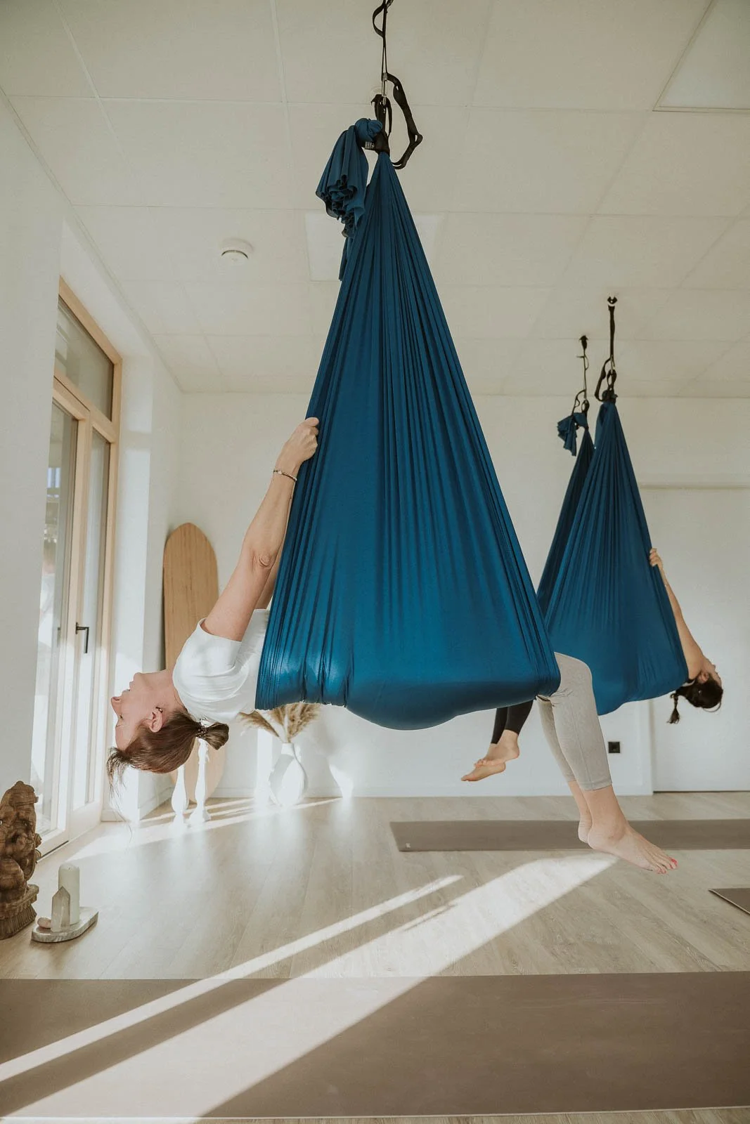 Zwei Frauen üben Aerial Yoga in Azure Sling-Hängematten im Yoga-Studio, blicken nach oben, entspannen sich, auf Holzboden, Sonnenlicht, weiße Wände.
