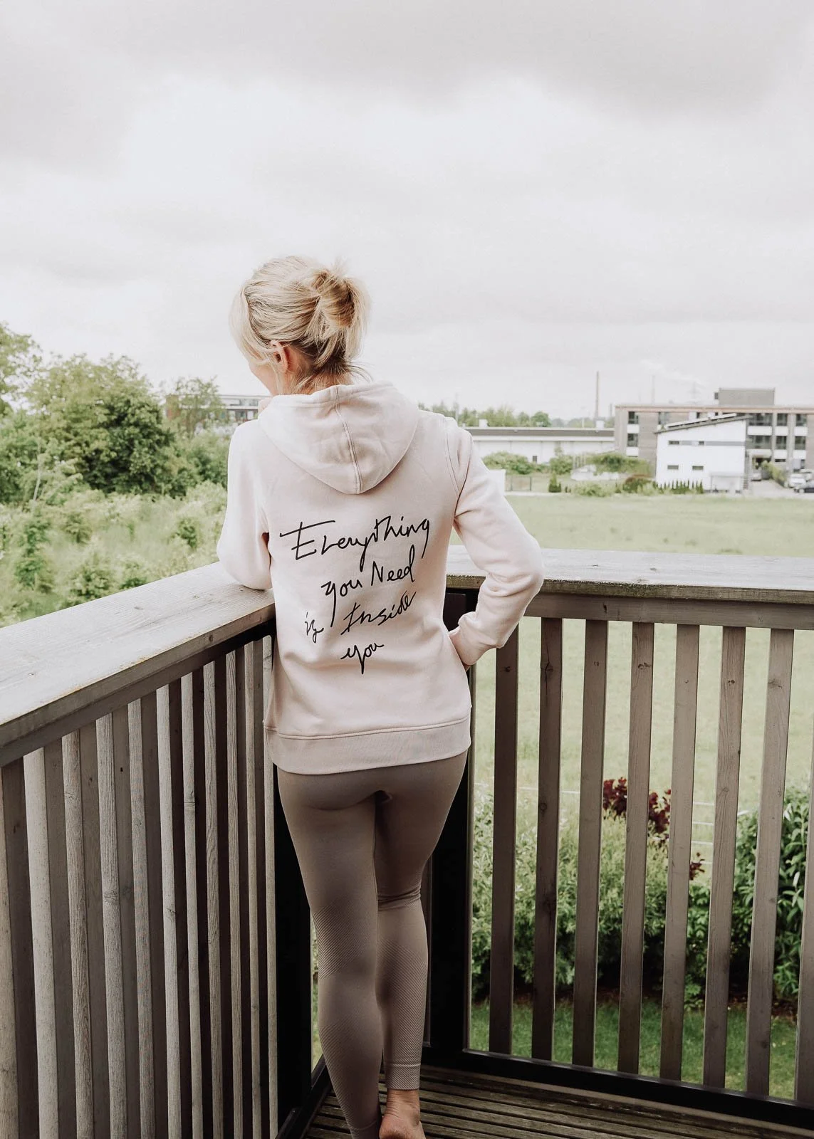 Frau steht auf einem Balkon, schaut auf die grüne Landschaft, trägt einen weißen Hoodie mit der Aufschrift ‚Everything you need is inside you‘, und trägt beige Leggings.