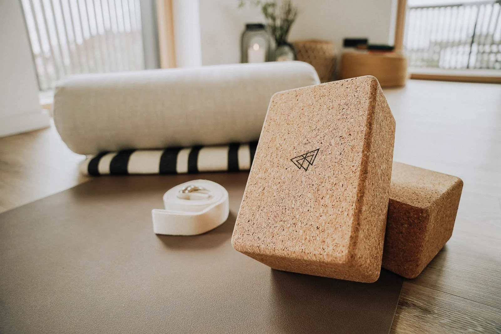 Yogamatte, Yogablock und Yogagurt auf einem Holzfußboden, im Hintergrund ein aufgerolltes Handtuch auf einer Couch in einem Wohnraum mit großen Fenstern und Balkon.