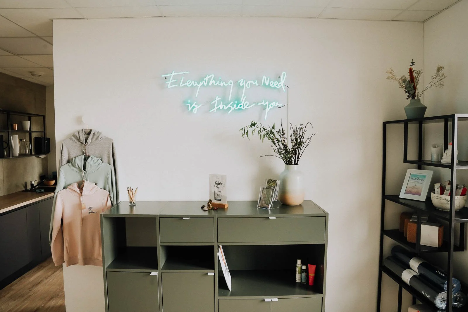 Weiße Wand mit Neon-Schildtext 'Everything you Need is Inside you' über einer grünen Kommode. Auf der Kommode stehen ein Vase mit Zweigen, Broschüren, ein Glas mit Stiften, kleine Figuren und ein Papier mit der Aufschrift 'Follow us'. An der linken S